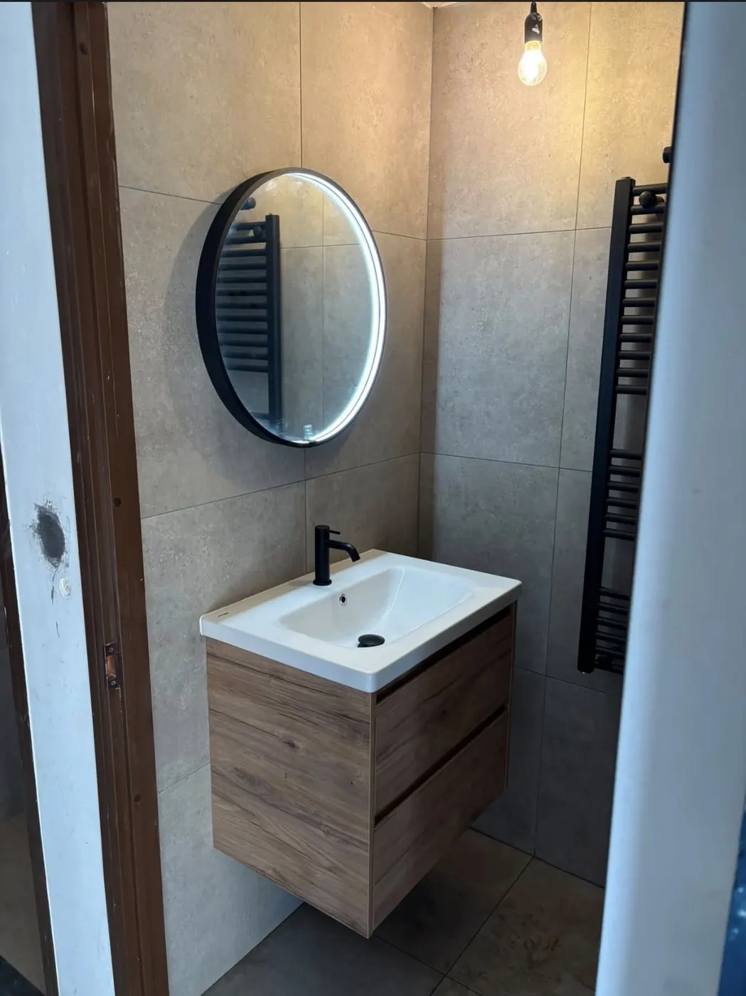 Badkamer in Wijchen met zwevend houten wastafelmeubel, ronde spiegel met verlichting en zwarte handdoekradiator